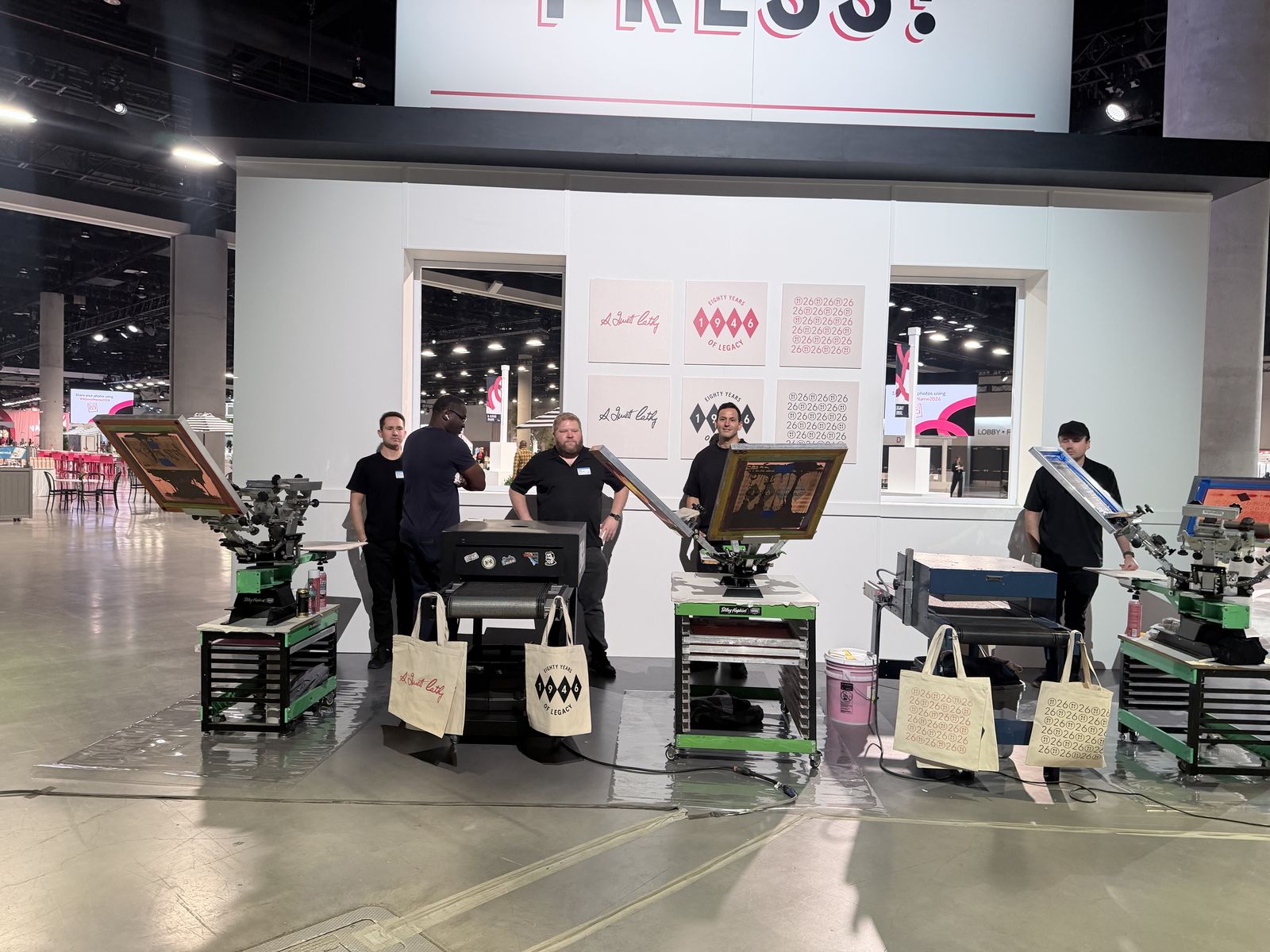 Multi-station live printing floor at a convention with presses, dryer, and printed tote bags