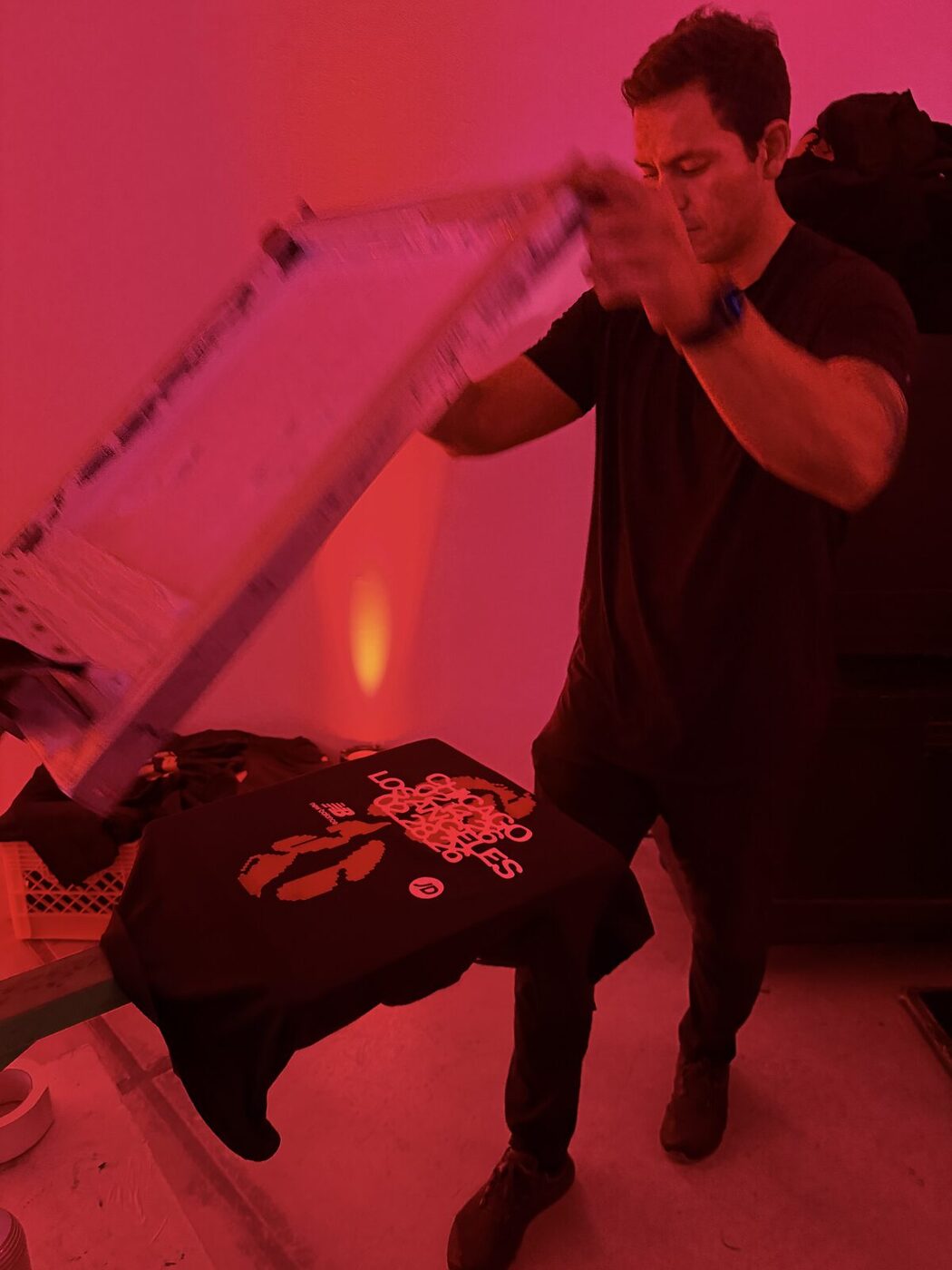 Printer lifting a screen to reveal a freshly printed shirt under red event lighting